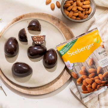 Rohe Mandel-Brownie-Schokoladeneier auf einem hölzernen Tisch mit Kakao, Datteln und Mandeln im Hintergrund.