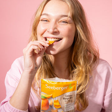 Eine Frau isst Seeberger Cashew-Mango-Mix vor rosafarbenem Hintergrund.
