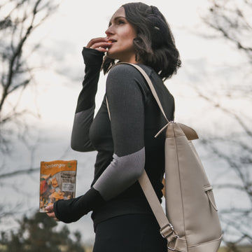 Frau isst Snack, hält Seeberger-Packung, trägt Rucksack, steht im herbstlichen Freien.